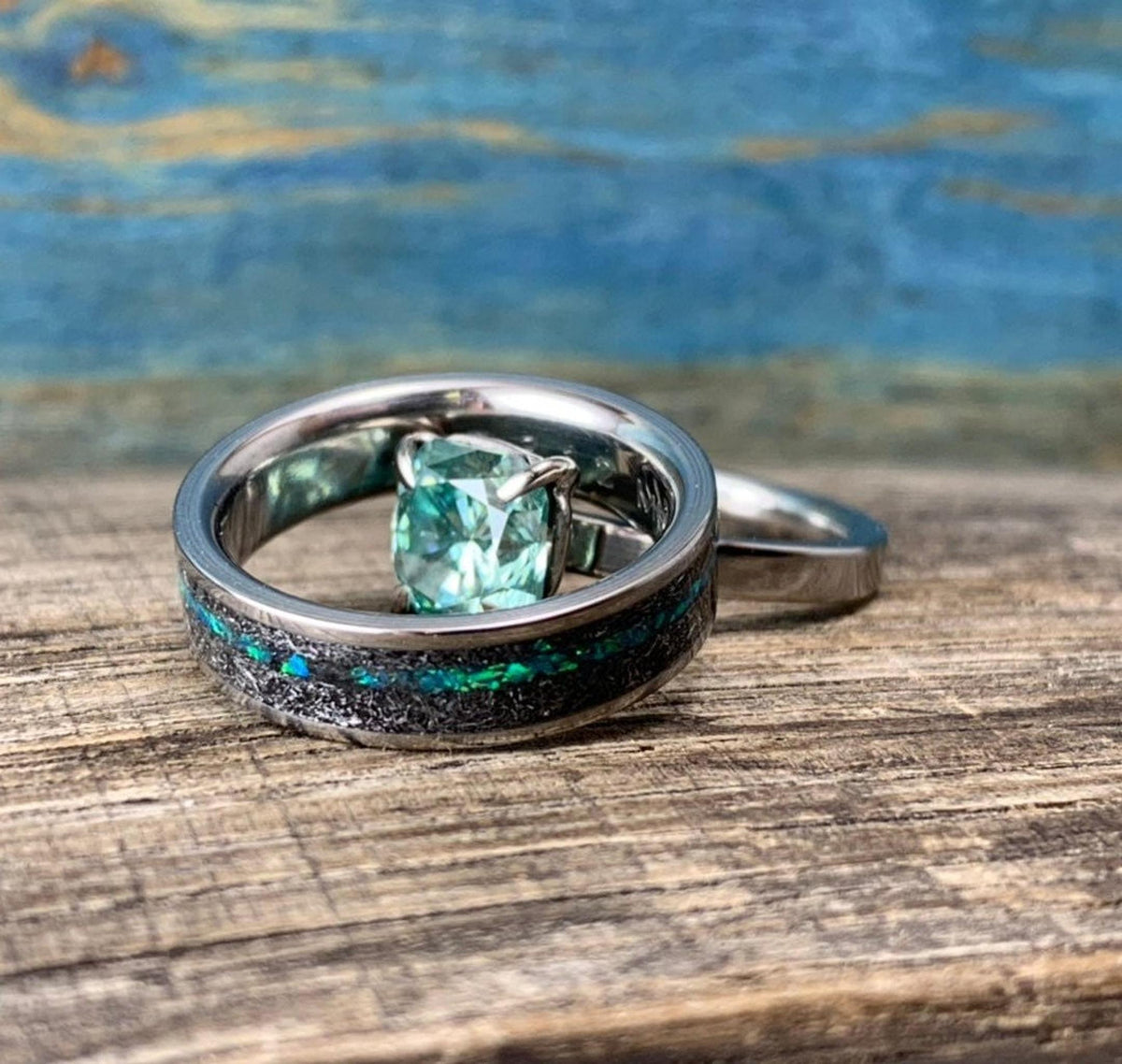 A silver-toned ring with a green square-cut stone is nestled inside a wider silver-toned band with a black and blue-green inlay, on a wooden surface.