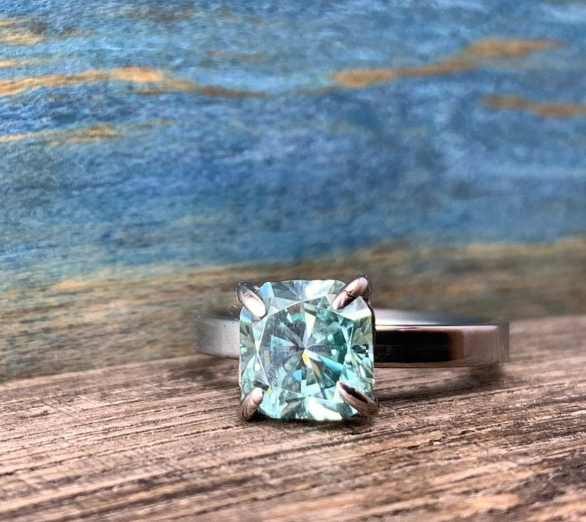 A silver-toned ring with a cushion-cut blue-green stone, set on a wooden surface with a blue and brown background.