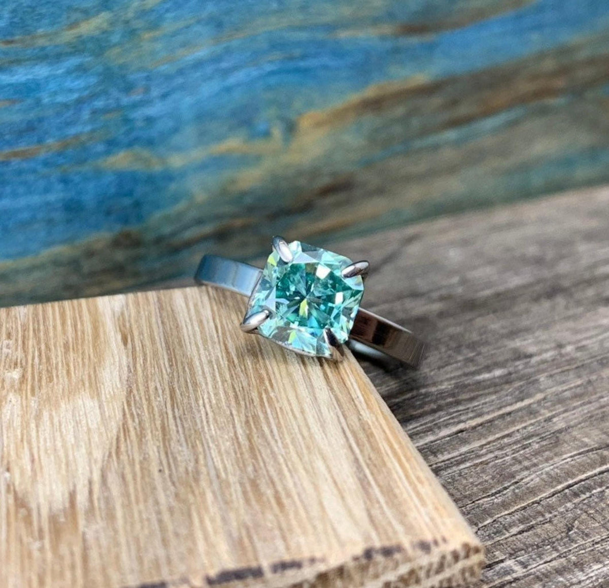 A silver-toned ring with a cushion-cut blue-green stone, angled on a light wooden block with a blue and brown background.