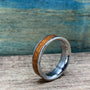 A silver-colored ring with a central brown wood inlay is angled on a wooden surface, with a blue-green background.