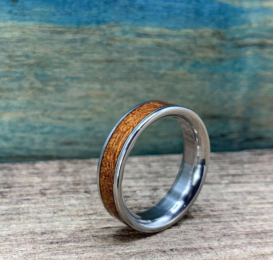 A silver-colored ring with a central brown wood inlay is angled on a wooden surface, with a blue-green background.