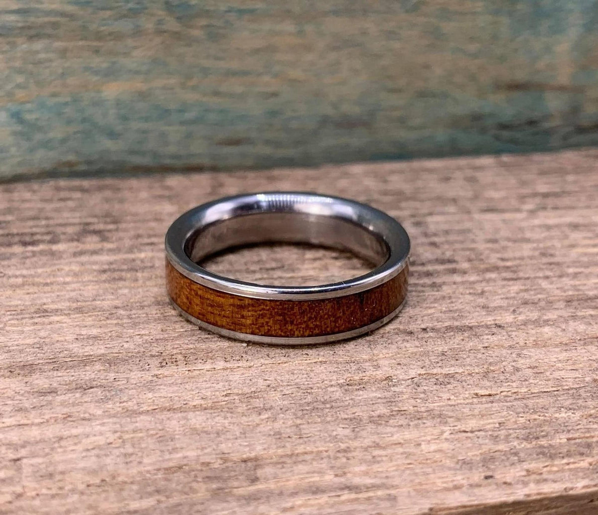 A silver-colored ring with a brown wood inlay is centered on a wooden surface, with a blue-green background.