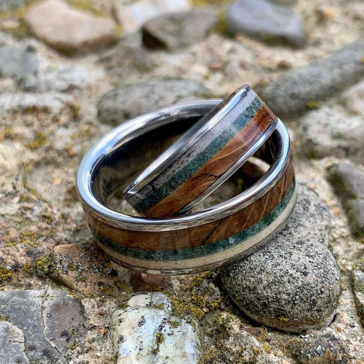 Two silver rings with wood, green, and cream inlay, one wider than the other, nested together on a rocky surface.