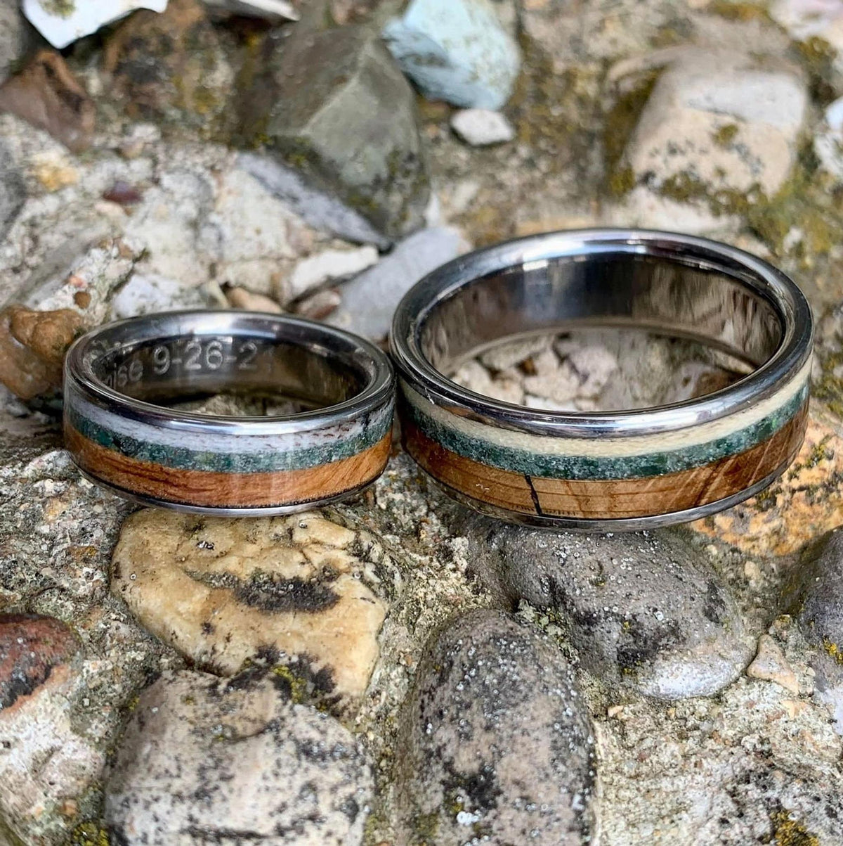 Two silver rings with wood, green, and cream inlay, one wider than the other, resting side-by-side on a rocky surface.