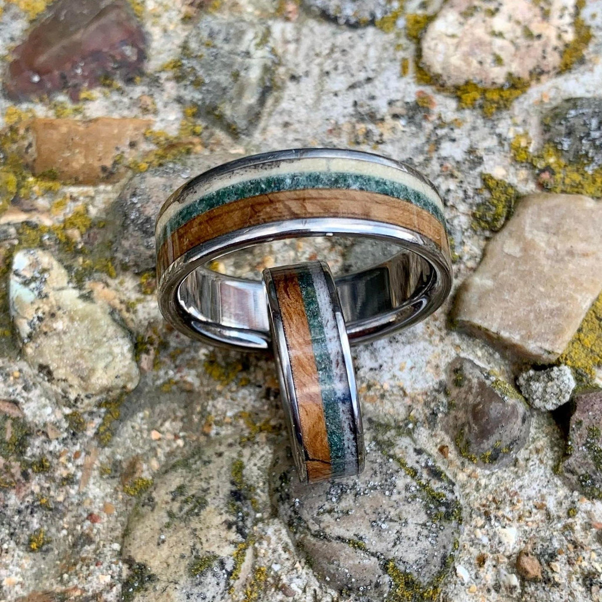 A wide silver ring with wood, green, and cream inlay, with a narrower matching ring leaning against it, on a rocky surface.
