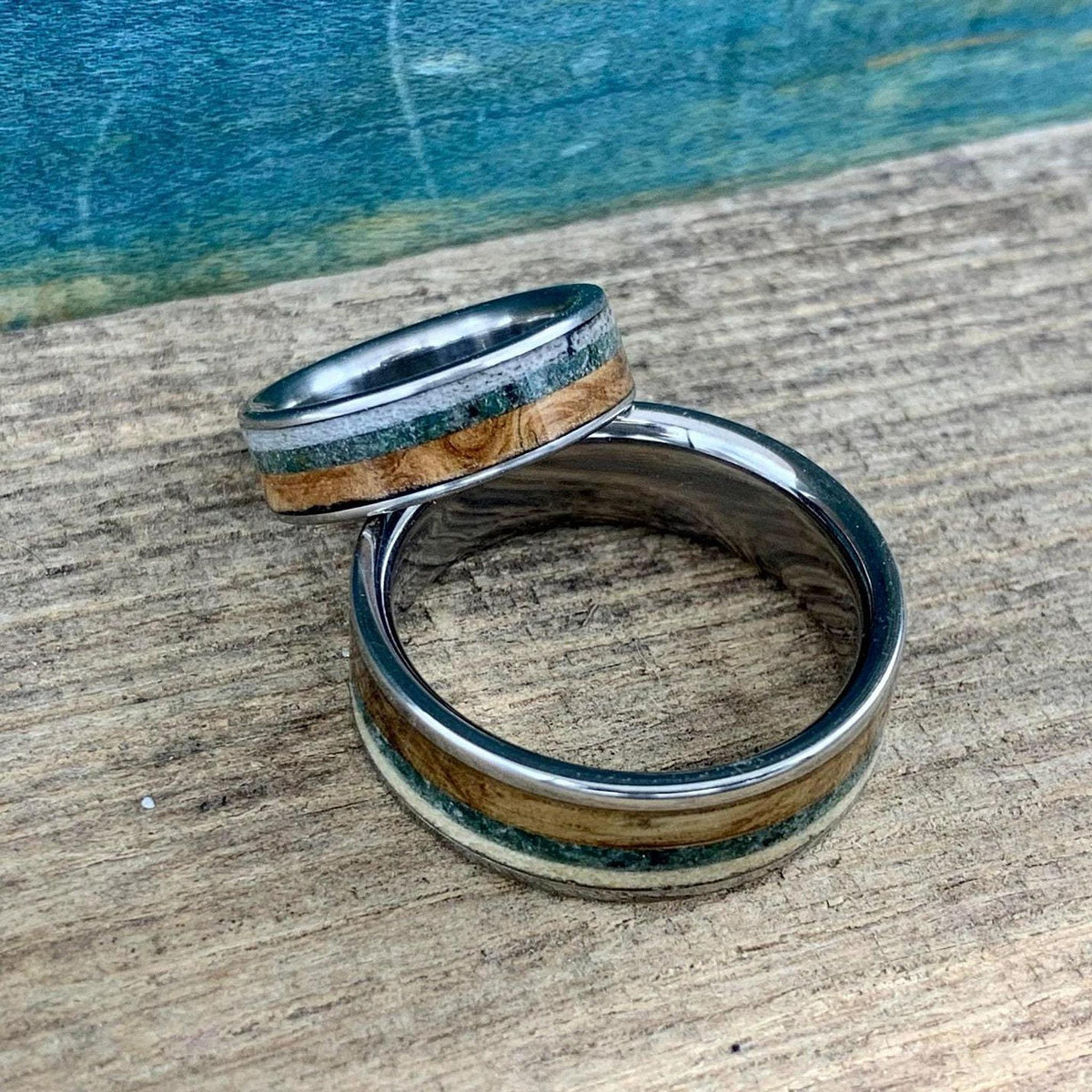 Two silver rings with wood, green, and cream inlay, one wider than the other, resting on a wooden surface with a blue background.