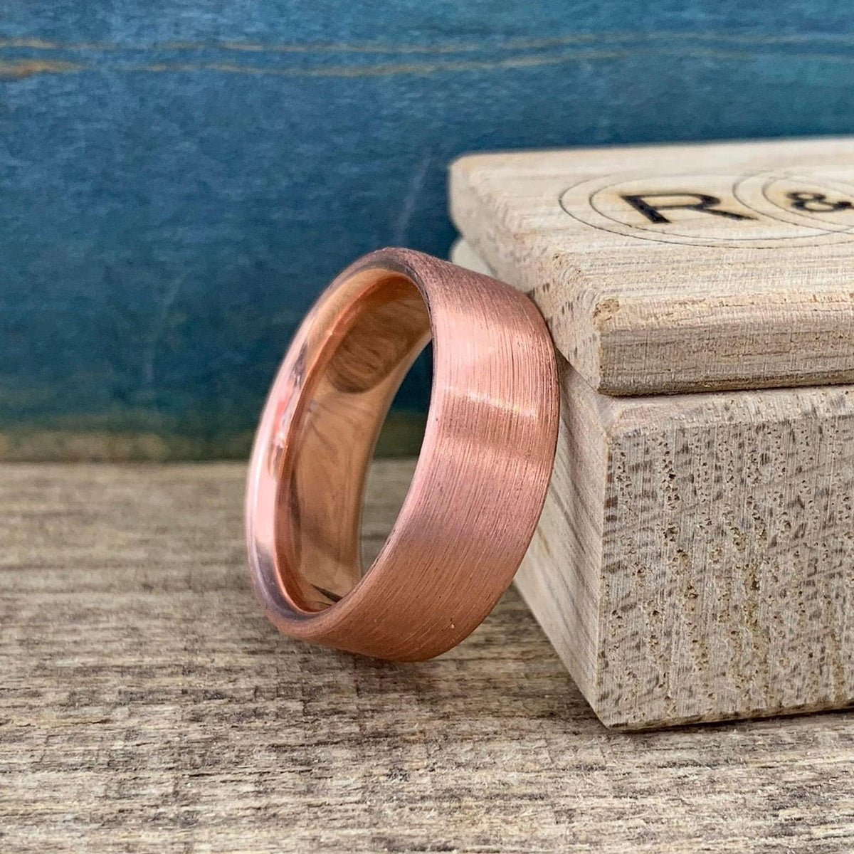 A brushed copper ring with a rounded interior, angled on a wooden surface next to a light wooden box.