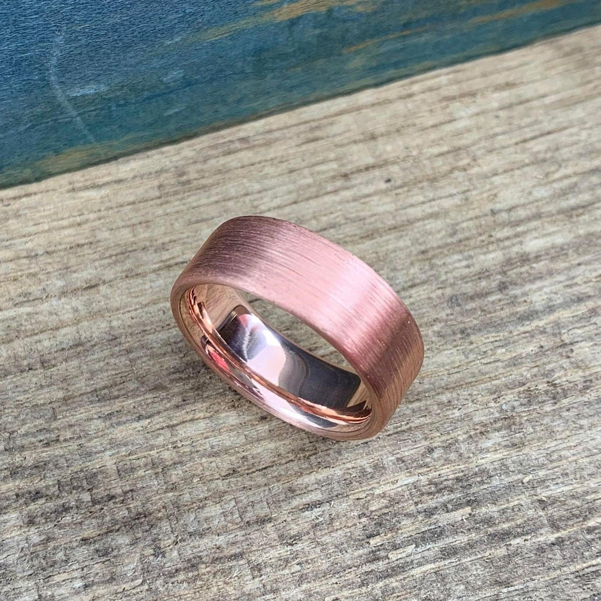 A brushed copper ring with a rounded interior, resting on a wooden surface with a blue background.