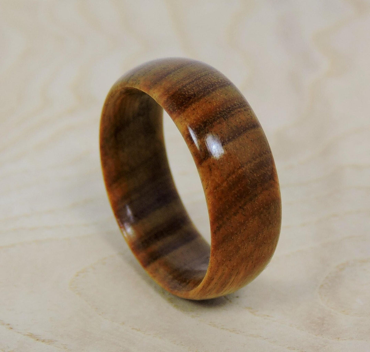 A smooth, polished, light brown wooden ring with darker brown horizontal grain lines stands upright on a light wooden surface.