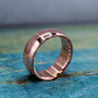 A rose gold-toned ring with a brushed finish on the outer band and a smooth, reflective inner band, resting on a distressed blue-green wooden surface.