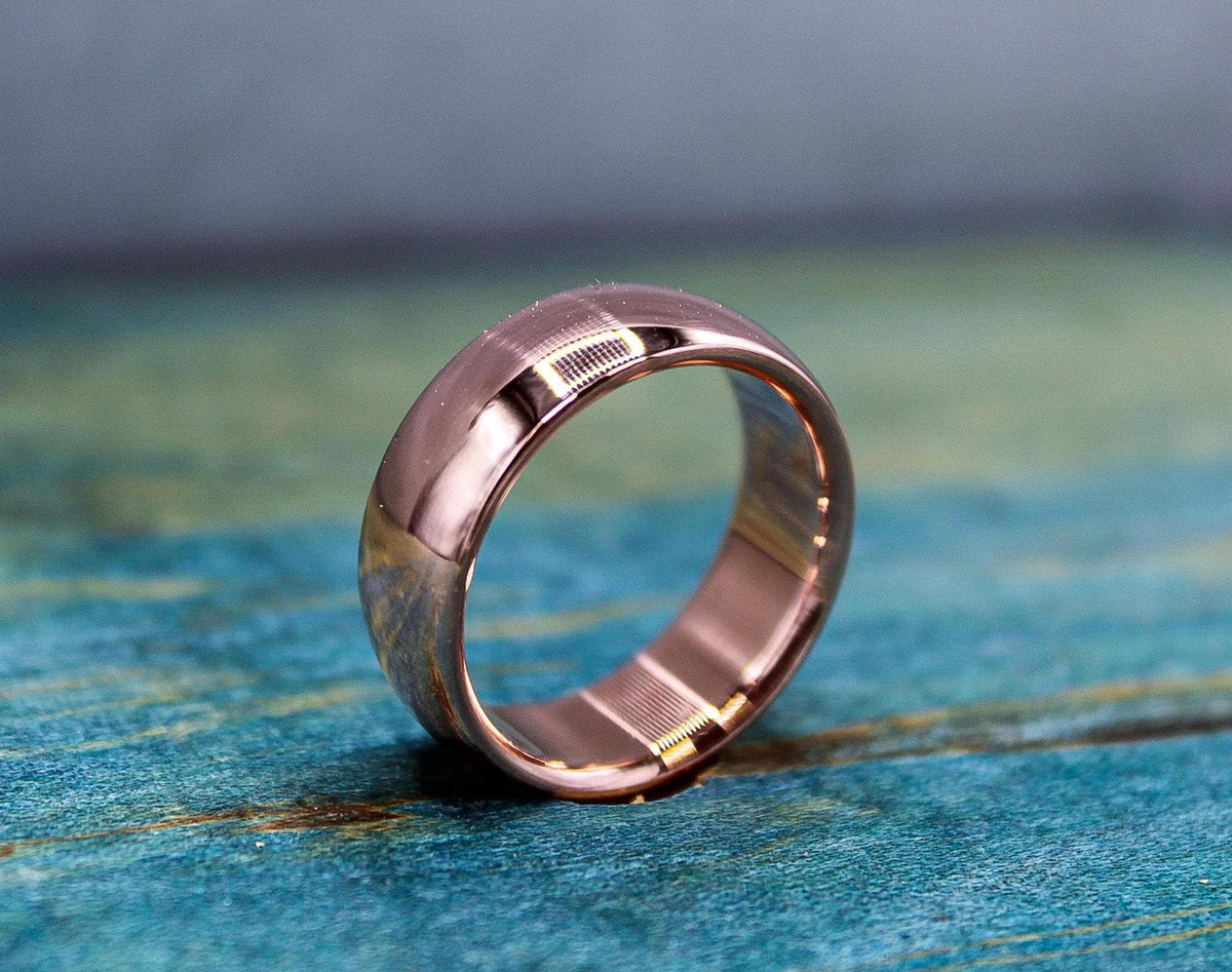 A rose gold-toned ring with a brushed finish on the outer band and a smooth, reflective inner band, resting on a distressed blue-green wooden surface.
