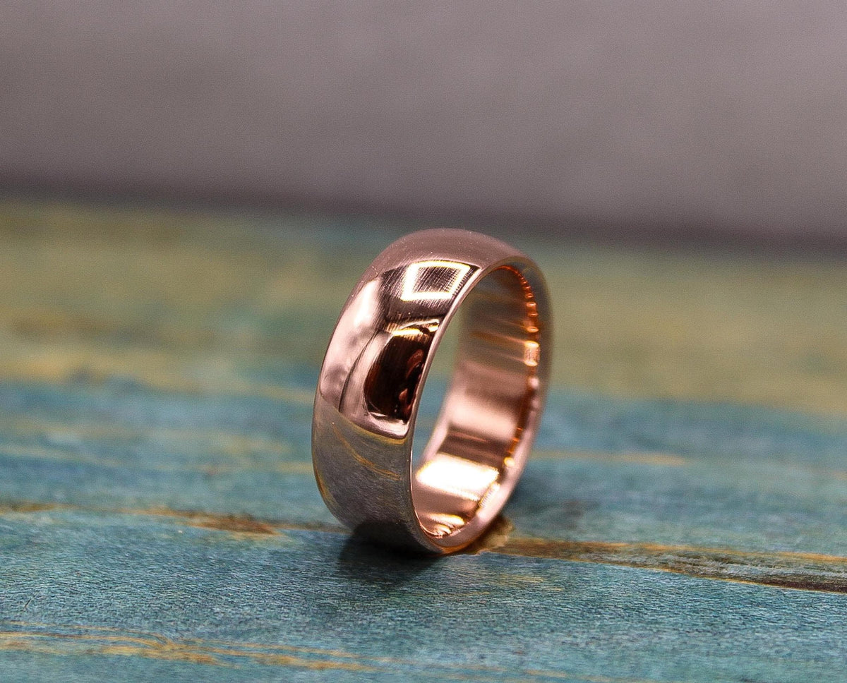A polished, rose gold-toned ring with a domed exterior and a flat interior, resting on a distressed blue-green wooden surface.