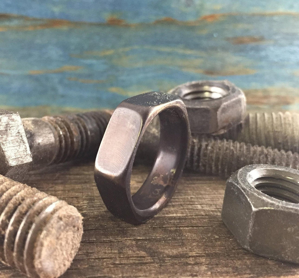 A dark hexagonal ring rests on a wooden surface surrounded by three large, rusty metal nuts and bolts.