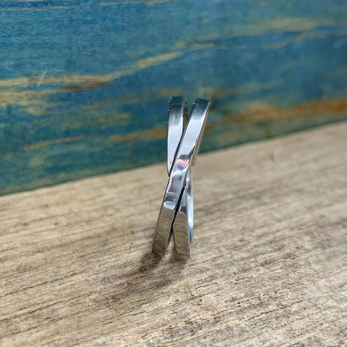 A shiny silver-colored crossover ring with two bands is shown from a side view, standing upright on a light wooden surface.