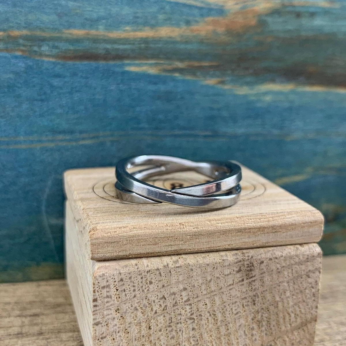 A shiny silver-colored crossover ring with two bands is displayed on a light wooden block, against a blue and brown background.