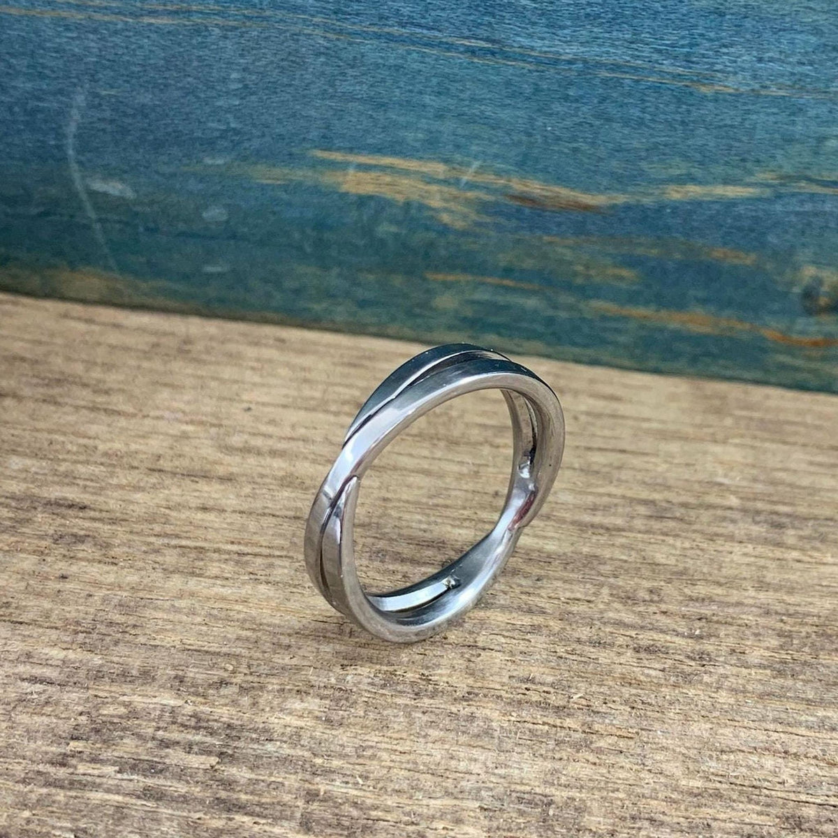 A shiny silver-colored crossover ring with two bands is displayed on a light wooden surface, viewed from a low, slightly angled perspective.