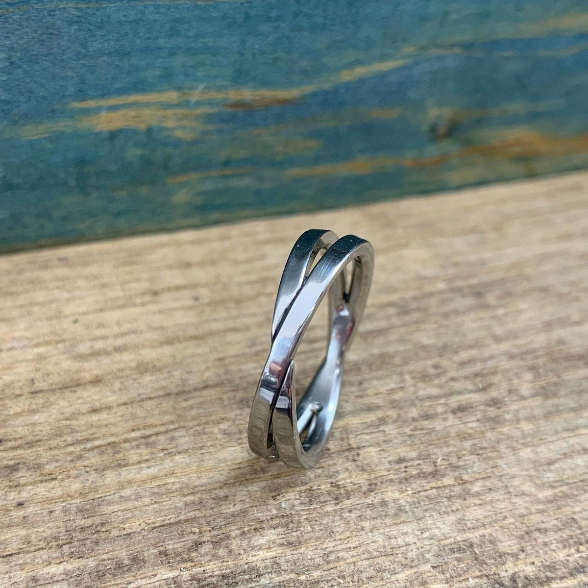 A shiny silver-colored crossover ring with two bands is displayed on a light wooden surface, viewed from a slight angle.