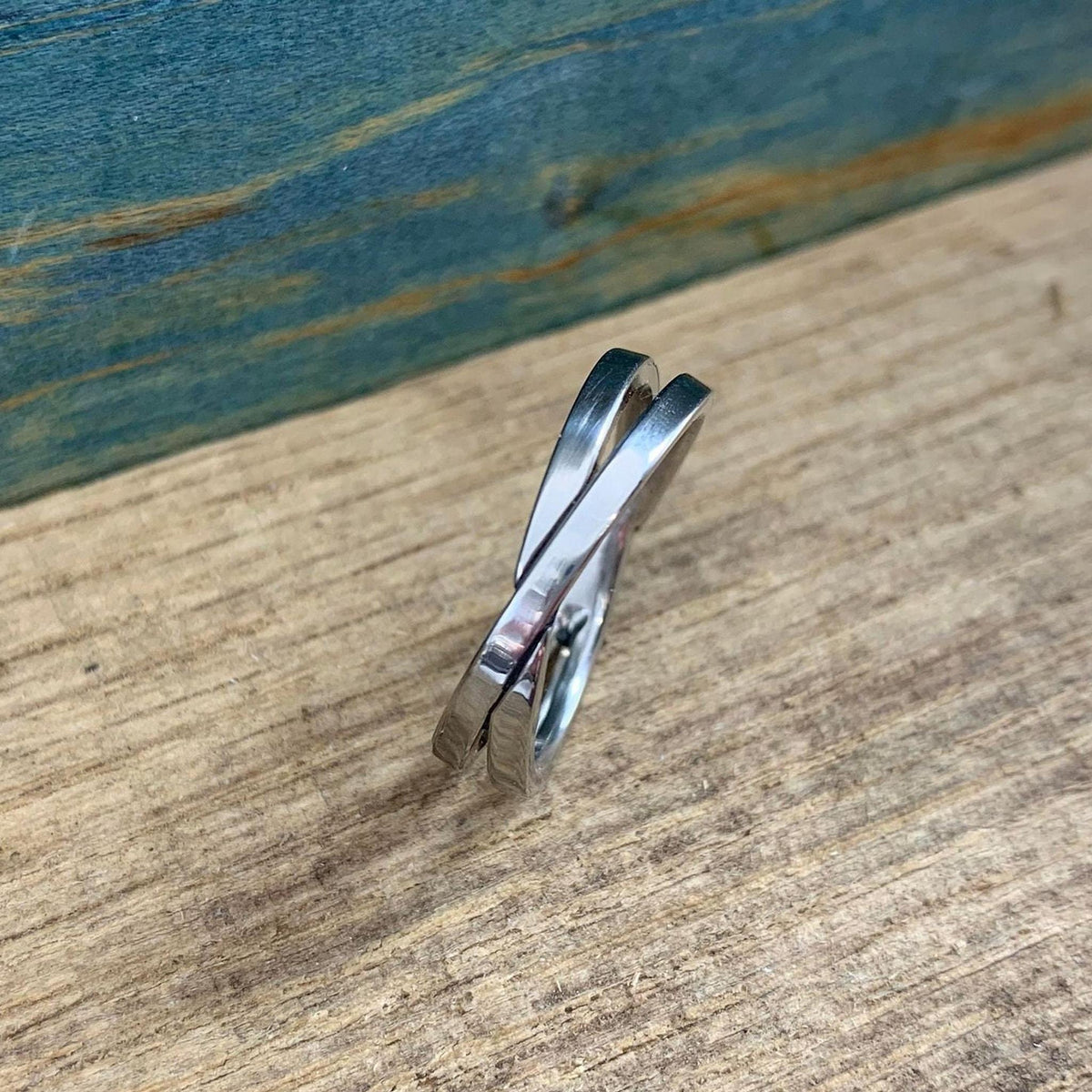 A shiny silver-colored crossover ring with two bands is shown from a side view, resting on a light wooden surface.