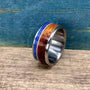 A silver-colored ring with a central dark wood inlay, a thin blue lapis lazuli inlay, and a guitar string inlay, on a light wood surface.