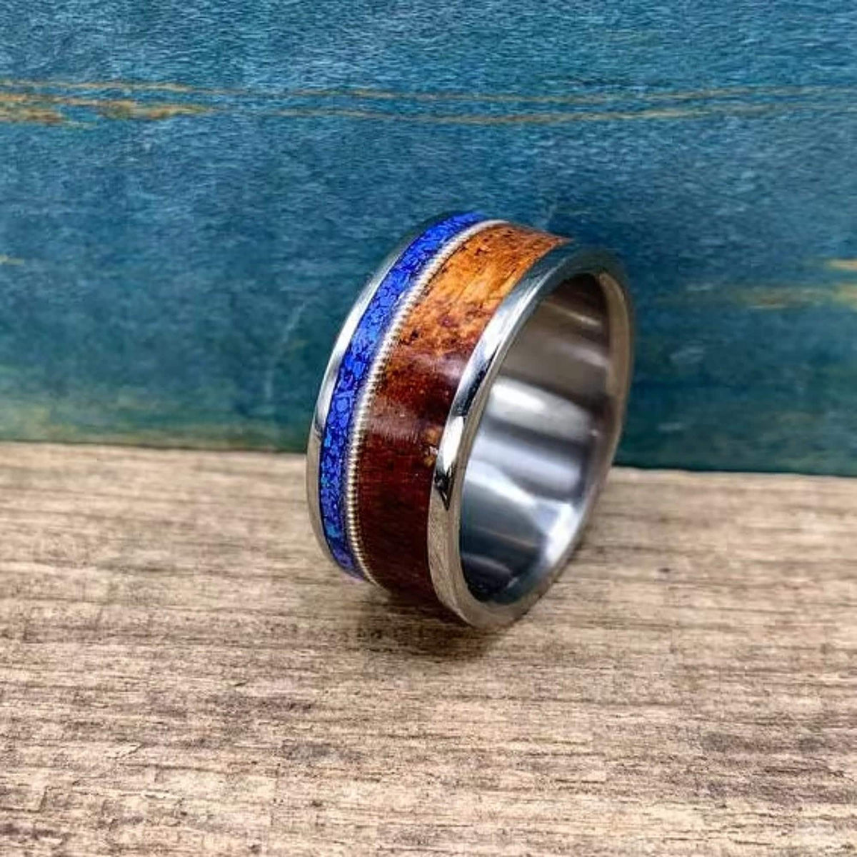 A silver-colored ring with a central dark wood inlay, a thin blue lapis lazuli inlay, and a guitar string inlay, on a light wood surface.