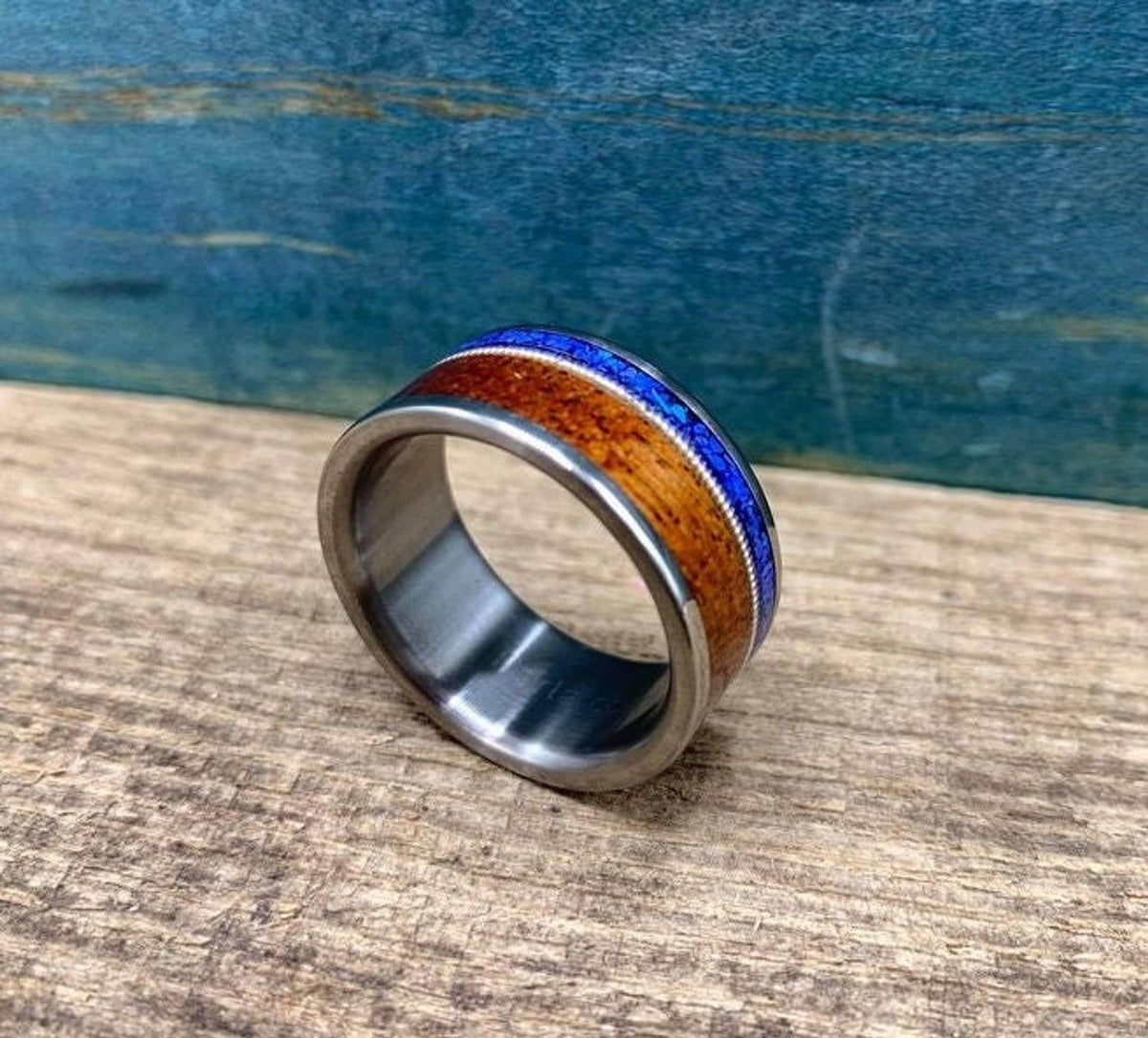 A silver-colored ring with a central dark wood inlay, a thin blue lapis lazuli inlay, and a guitar string inlay, on a light wood surface.