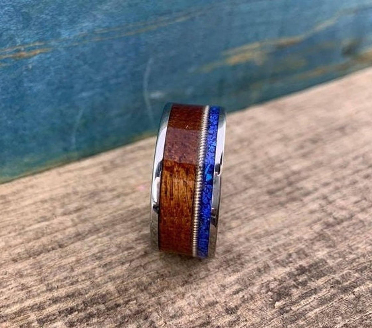 A silver-colored ring with a central dark wood inlay, a thin blue lapis lazuli inlay, and a guitar string inlay, on a light wood surface.