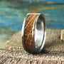 A silver-colored ring with a light brown wood inlay, featuring horizontal grain lines, rests on a textured gray surface with a blue and green background.