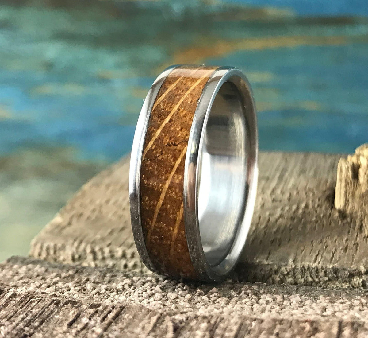 A silver-colored ring with a light brown wood inlay, featuring horizontal grain lines, rests on a textured gray surface with a blue and green background.