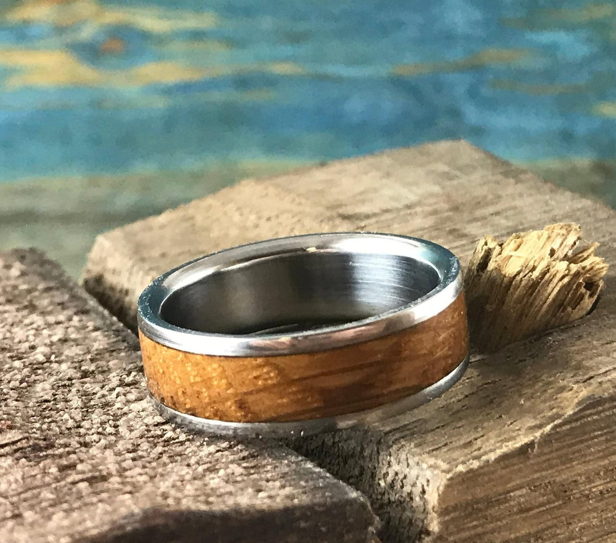 A silver-colored ring with a light brown wood inlay rests on a light brown wooden block next to a small piece of natural wood, with a blue and green background.