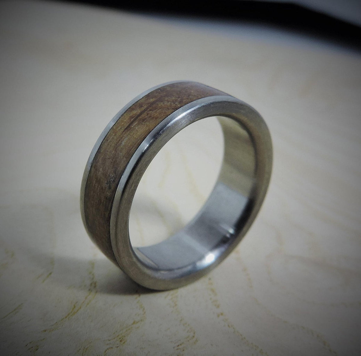 A silver-colored ring with a smooth, light brown wood inlay is angled on a light wooden surface.