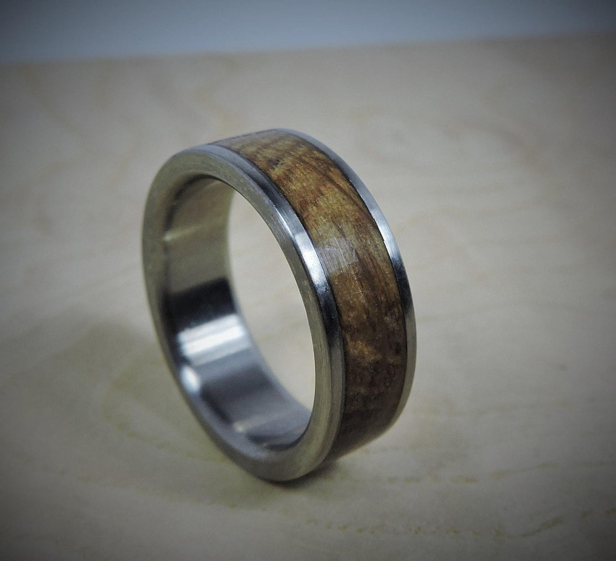 A silver-colored ring with a light brown wood inlay, showing subtle grain, is angled on a light wooden surface.