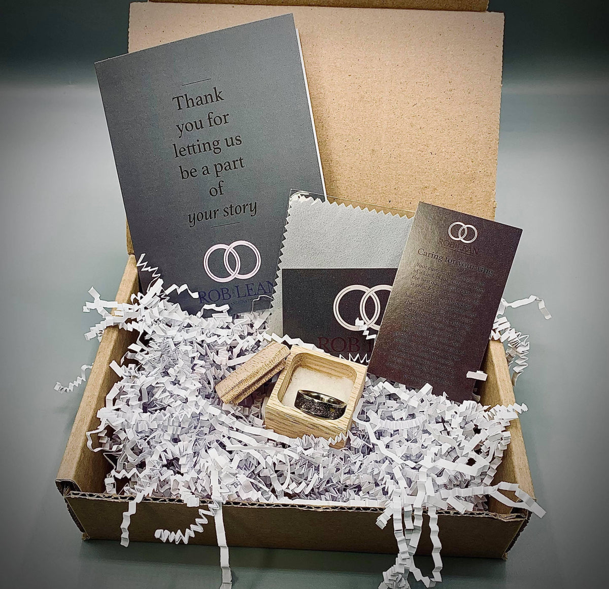 An open cardboard box contains a ring in a small wooden box, shredded white paper, and two black cards with white text and logos.