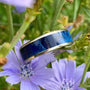 A silver-colored titanium ring with a blue box elder wood inlay is nestled among purple chicory flowers and green leaves.
