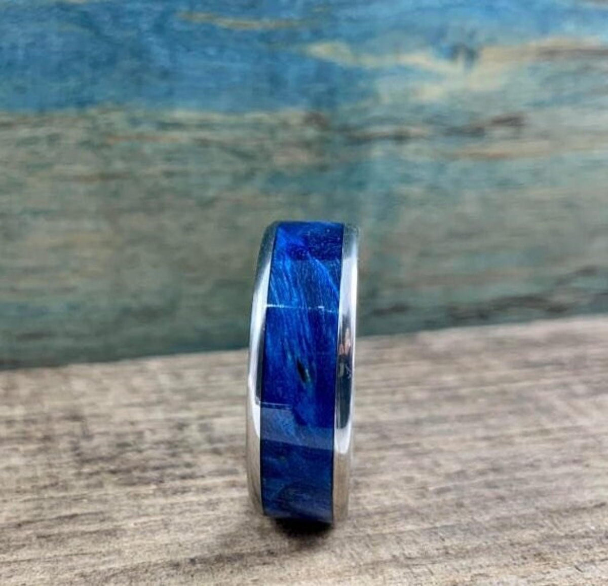 A silver-colored titanium ring with a blue box elder wood inlay is shown from a front-facing perspective, on a blurred wooden surface.