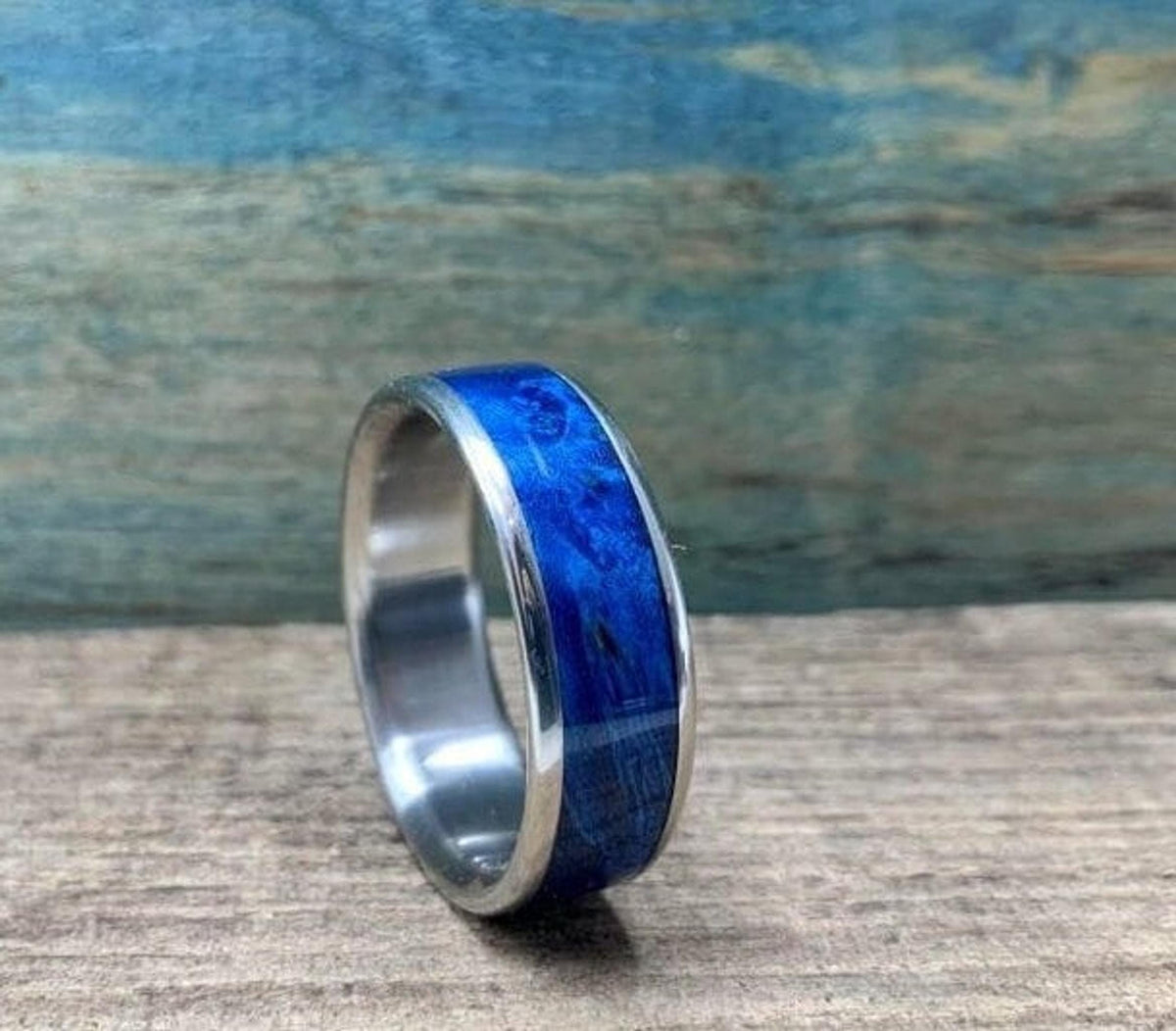 A silver-colored titanium ring with a blue box elder wood inlay is angled on a light brown wooden surface, with a blurred blue wooden background.