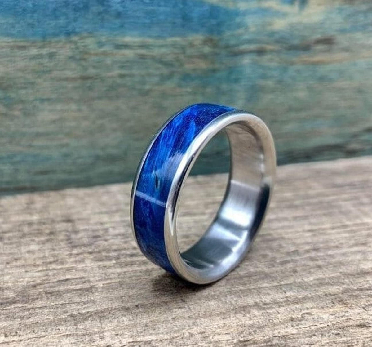 A silver-colored titanium ring with a blue box elder wood inlay is angled on a light brown wooden surface, with a blurred blue wooden background.