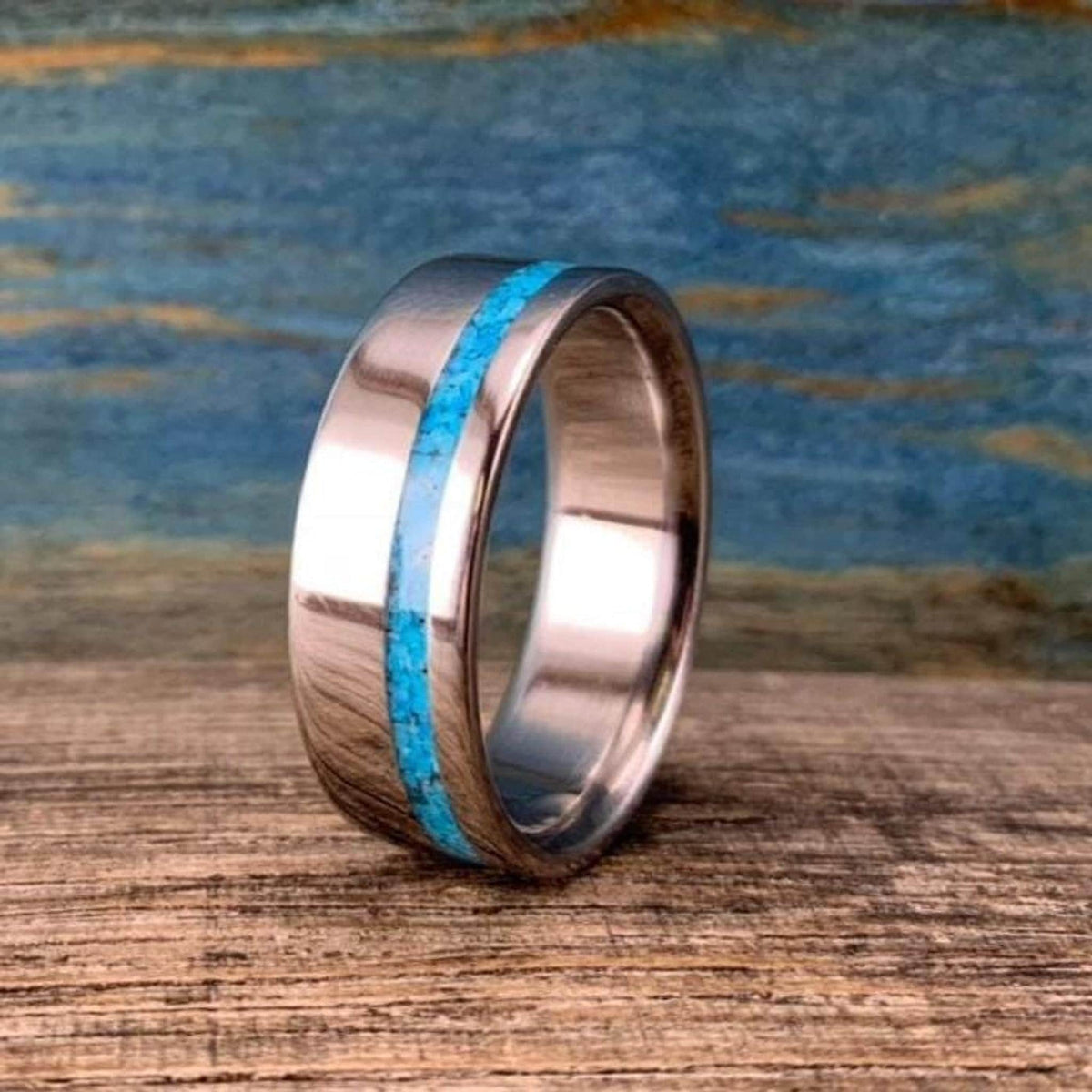 A silver-colored ring with a thin turquoise inlay band, angled on a wooden surface with a blue background.