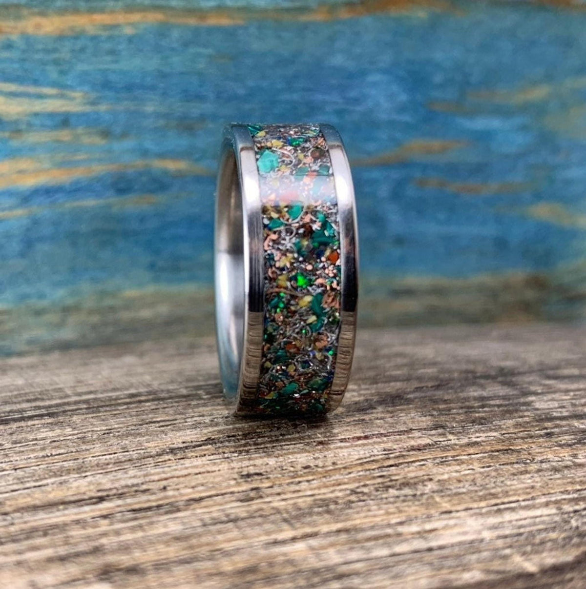 A silver-colored ring with a colorful inlay of green, orange, and brown fragments, seen from the side on a wooden surface.