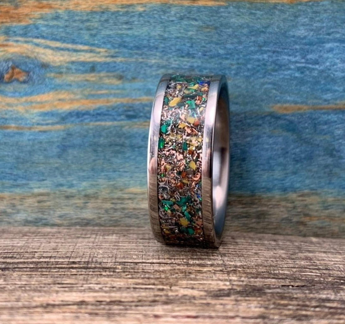 A silver-colored ring with a colorful inlay of green, orange, and brown fragments, viewed from the side on a wooden surface.
