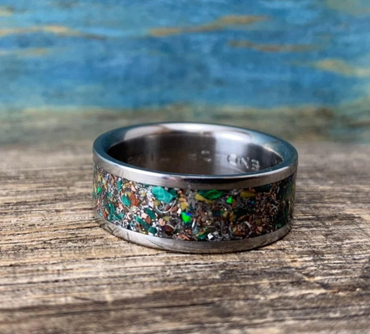 A silver-colored ring with a colorful inlay of green, orange, and brown fragments, with text visible on the inside band.