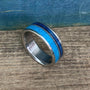 A silver-colored ring with a wide turquoise inlay band, a thin dark blue inlay, and a thin silver-colored textured band, on a wooden surface.