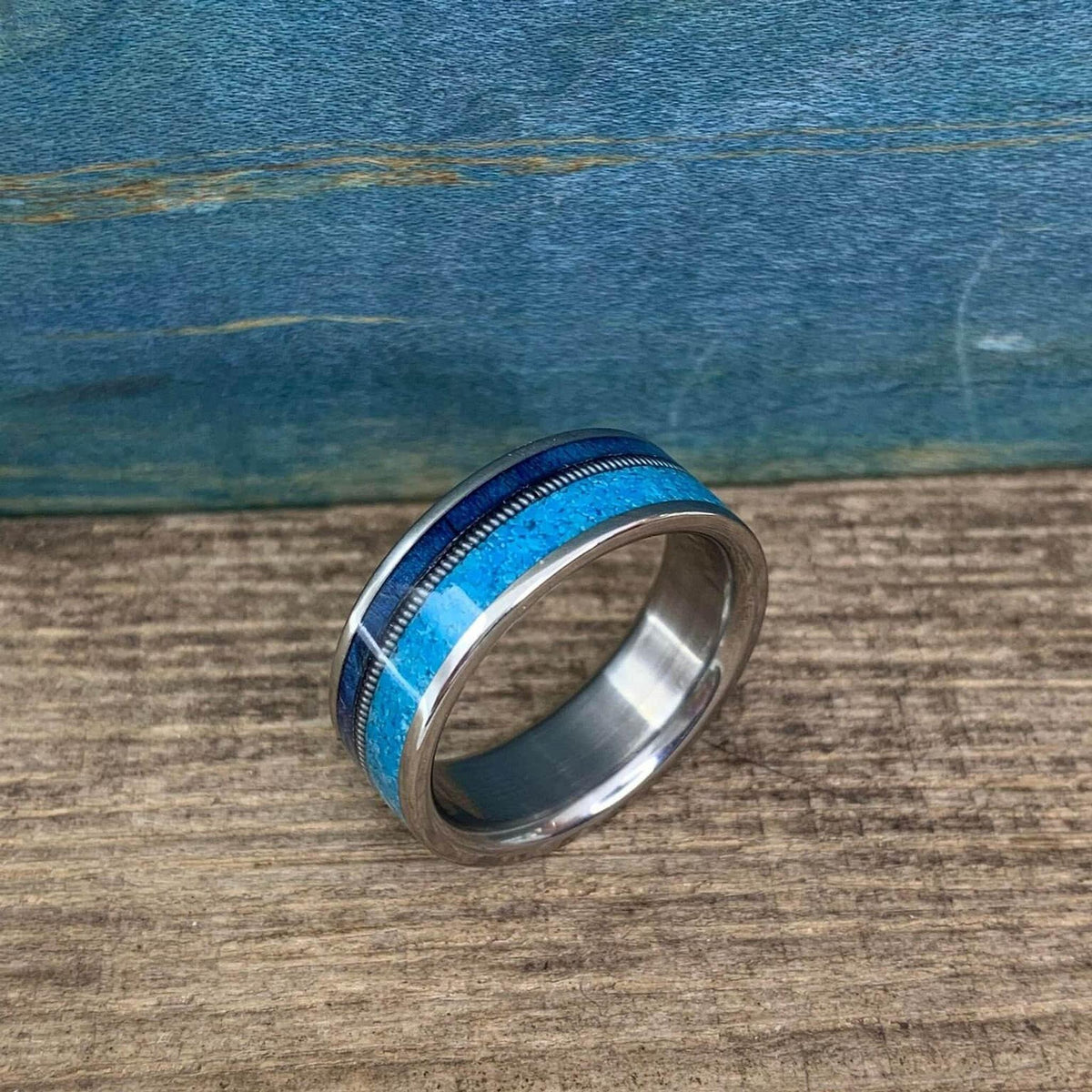 A silver-colored ring with a wide turquoise inlay band, a thin dark blue inlay, and a thin silver-colored textured band, on a wooden surface.