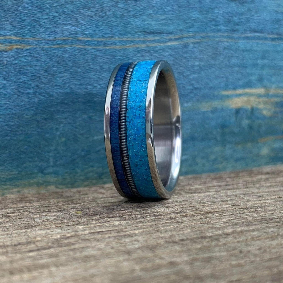 A silver-colored ring with a wide turquoise inlay band, a thin dark blue inlay, and a thin silver-colored textured band, on a wooden surface.