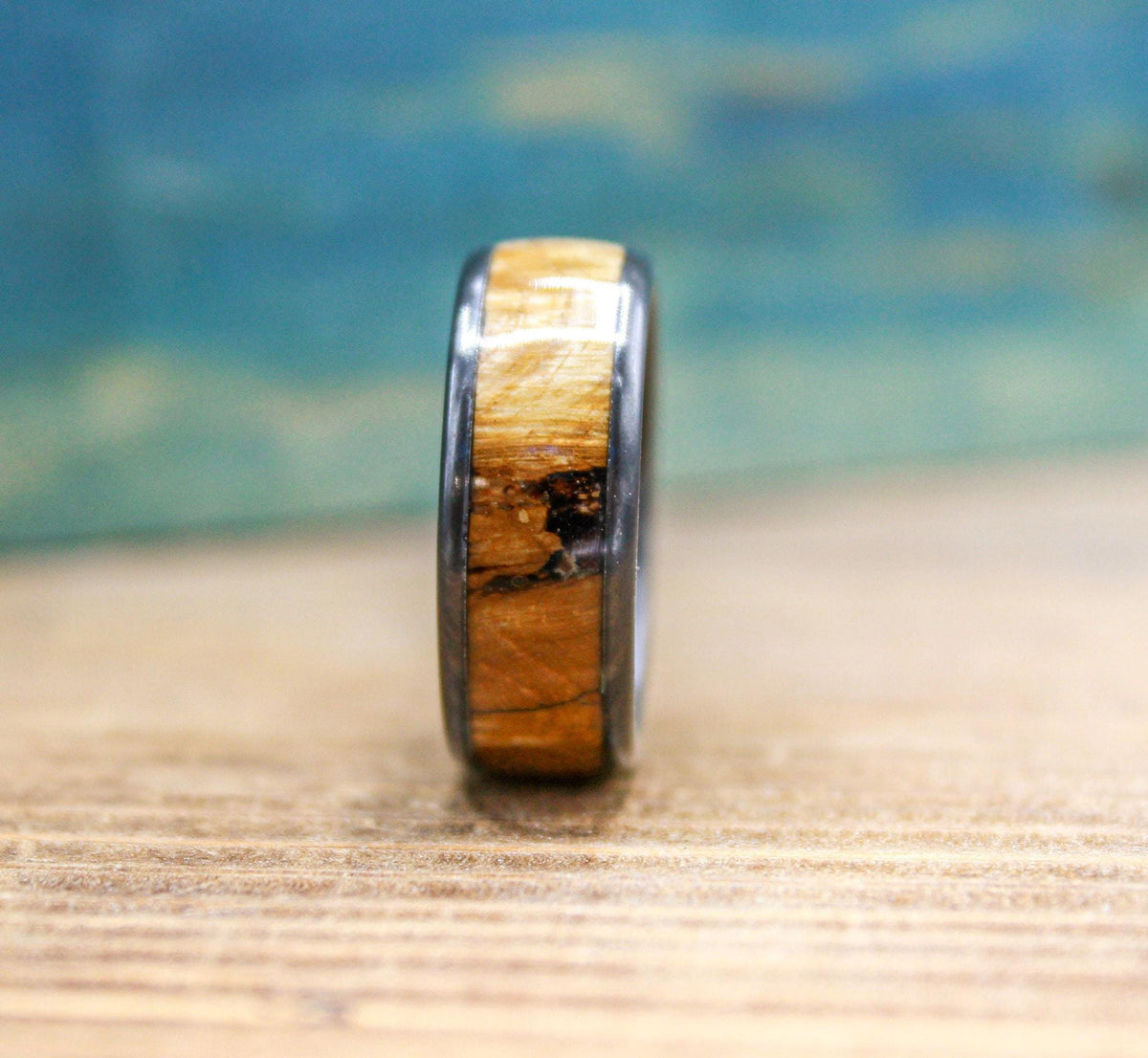 A black titanium ring with a light brown wood inlay is shown from a side view, highlighting the wood grain and black edges.