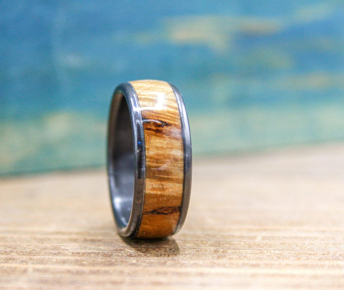 A black titanium ring with a light brown wood inlay, displaying varied wood patterns, is positioned on a wooden surface.