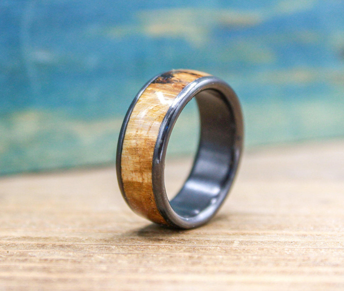 A black titanium ring with a light brown wood inlay, showing a smooth, polished surface, rests on a wooden table.
