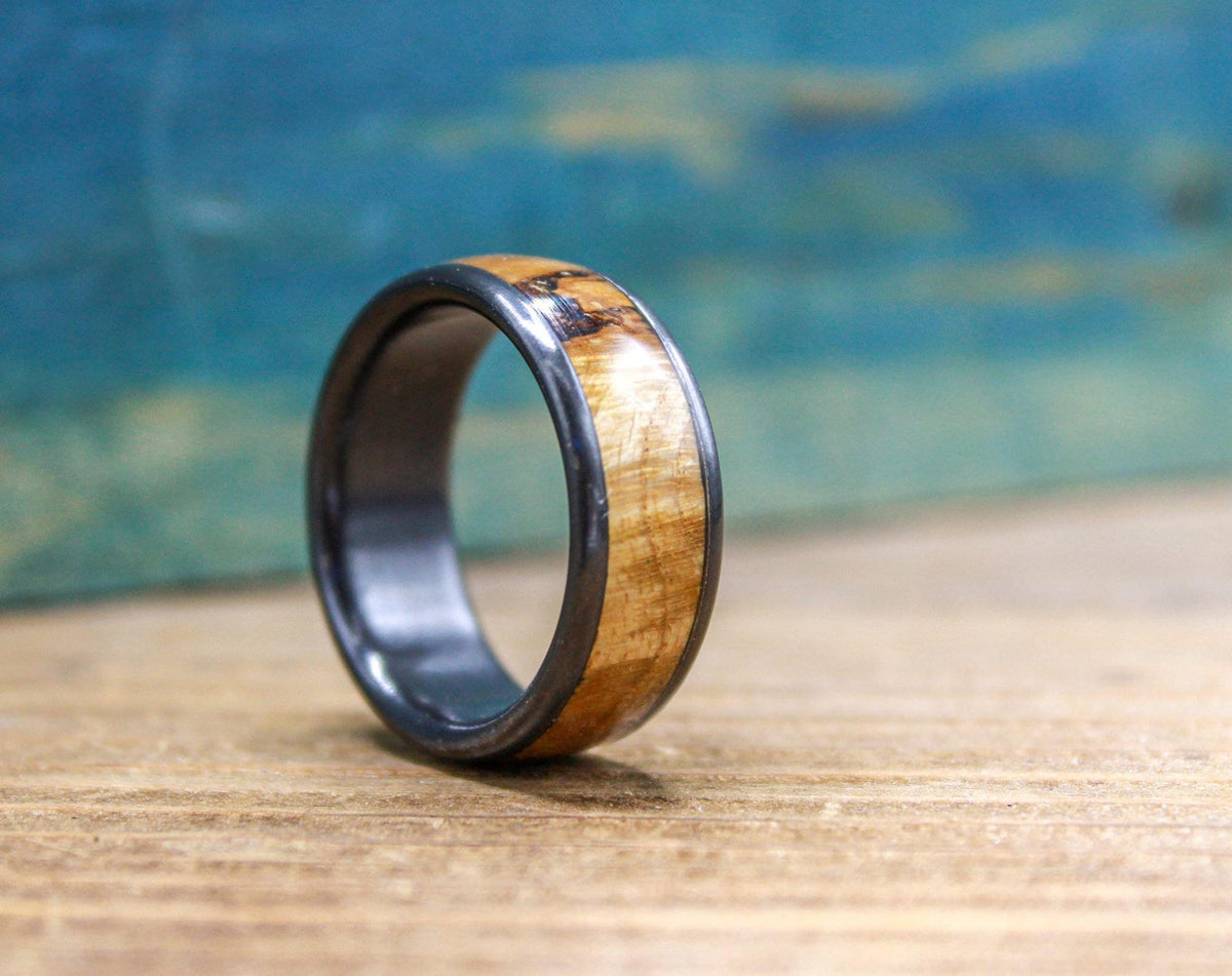 A black titanium ring with a light brown wood inlay, featuring natural wood markings, is displayed on a wooden background.