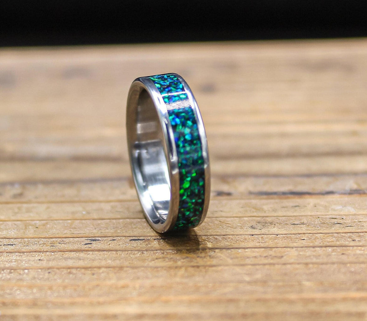 A silver-colored ring with a green and blue speckled inlay is angled on a wooden surface, with the inlay facing up.