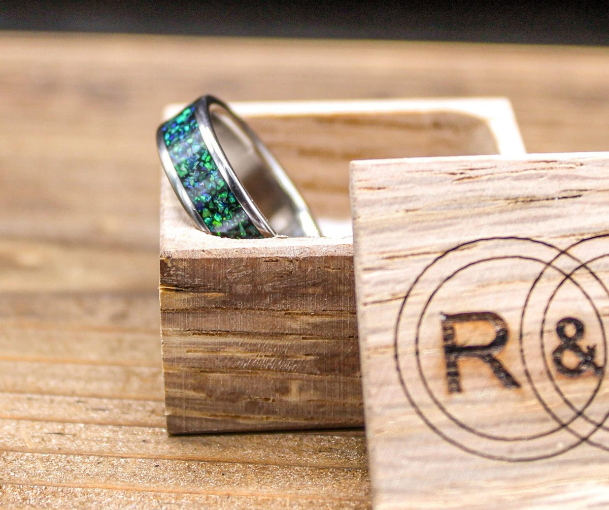 A silver-colored ring with a green and blue speckled inlay rests inside an open light wood box with a circular R&L logo.