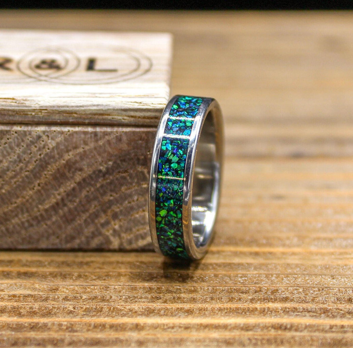 A silver-colored ring with a green and blue speckled inlay sits on a wooden surface next to an open light wood box with an R&L logo.